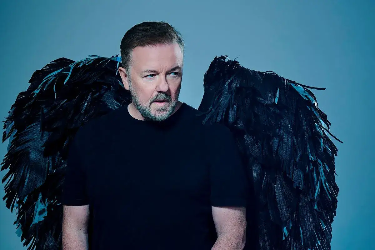 Ricky Gervais, z czarnymi skrzydłami anioła, gotowy na swój kolejny występ stand-up.