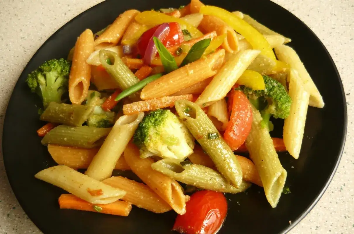 Kolorowy makaron penne z brokułami, marchewką, papryką i pomidorkami. Pyszny makaron z warzywami na czarnym talerzu.
