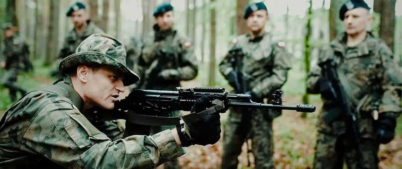 Żołnierz WOT w kamuflażu, celujący z karabinu w lesie. W tle inni żołnierze.