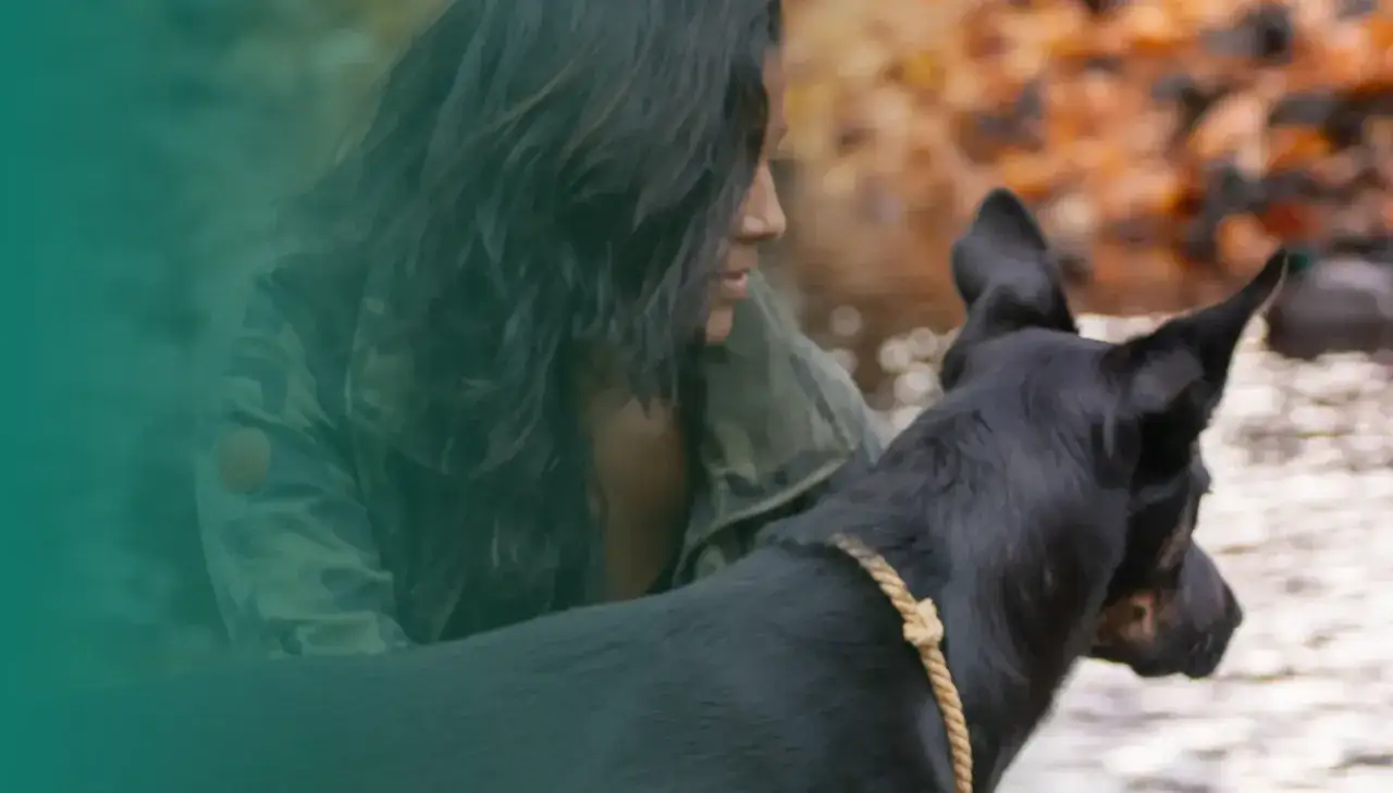 En kvinna med mörkt hår och en kamouflagejacka tittar på sin svarta hund. Deras ledarskap och band syns tydligt.