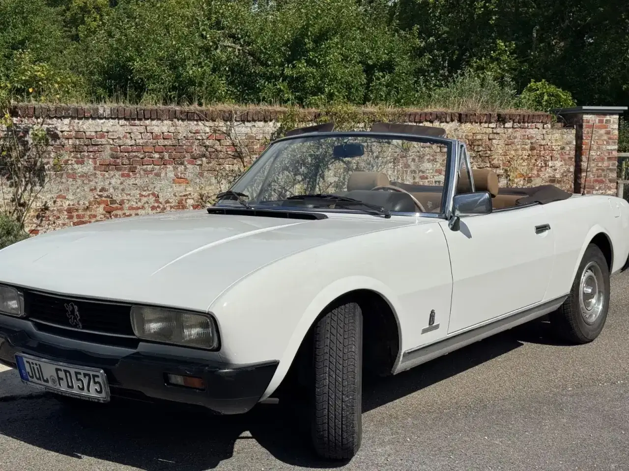 Biały kabriolet Peugeot 504 z brązowym wnętrzem, zaparkowany przy ceglanym murze.