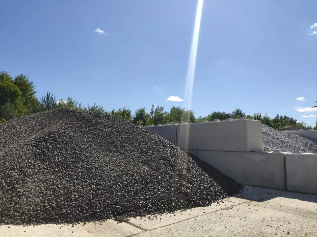Ogromna hałda ciemnego bazaltu na placu budowy, obok betonowych bloków i zielonych drzew pod błękitnym niebem.
