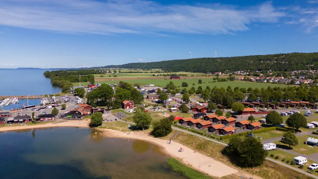 Gr&auml;nna camping vid V&auml;tterns strand med sm&aring; r&ouml;da stugor, b&aring;thamn, strand och gr&ouml;nskande omgivningar.