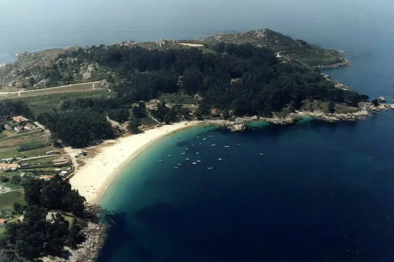 Vista aérea de **praia da Mourisca**, con arena dorada, aguas turquesas y barcos. Al fondo, colinas verdes y casas.