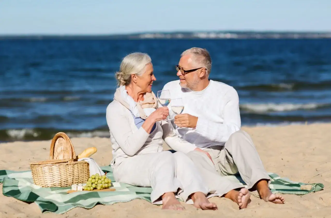 Uśmiechnięta para seniorów wznosi toast kieliszkami białego wina na plaży. Idealne mrzeżyno wczasy dla seniora.