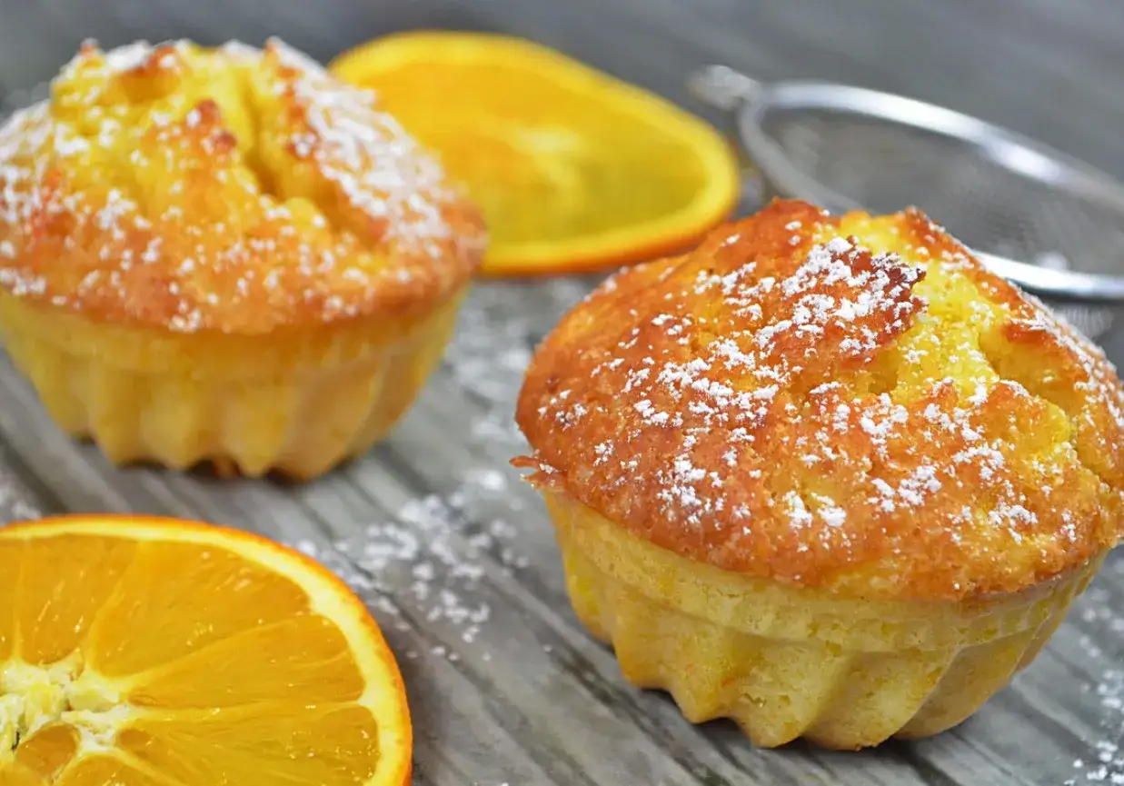 Puszyste muffinki pomarańczowe, idealne na przepis na wilgotne i puszyste muffinki. Posypane cukrem pudrem, obok plastry pomarańczy.