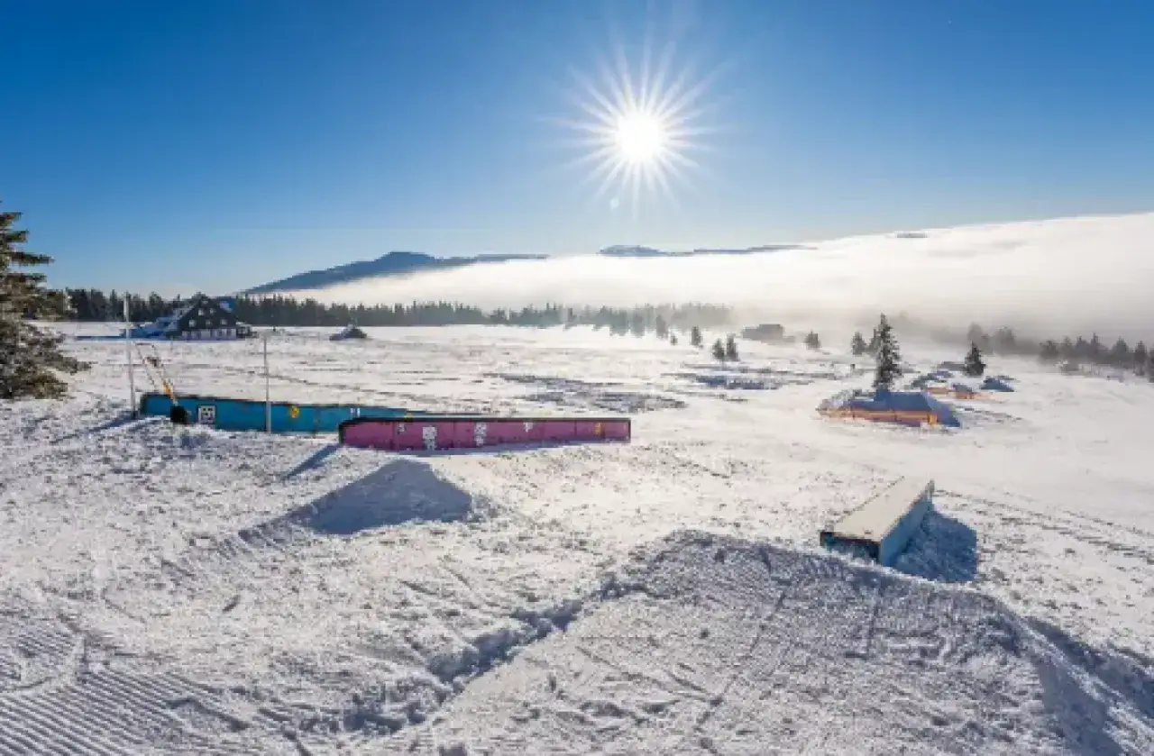 Słoneczny dzień w spindleruv mlyn snowpark. Widać kolorowe przeszkody i budynki w oddali, a poniżej morze chmur.