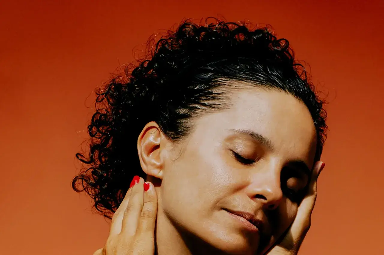 A woman with curly hair and closed eyes, her hand gently touching her face, embodies timeless beauty.