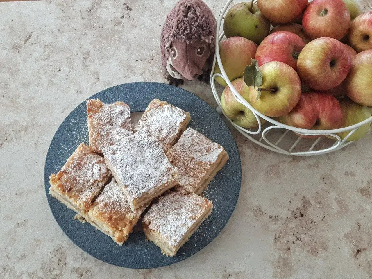 Pyszna, najlepsza szarlotka, która zawsze wychodzi, posypana cukrem pudrem. Obok koszyk z jabłkami i pluszowy jeż.