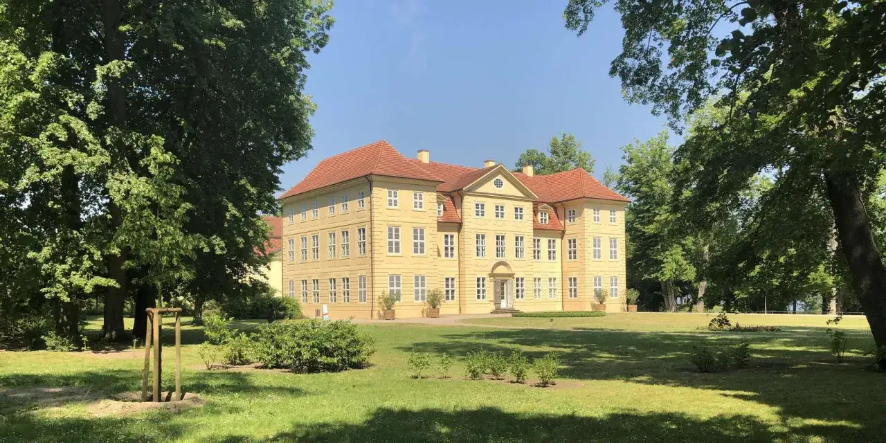 Das Schlossinsel Mirow, ein gelbes Barockschloss mit roten Dächern, steht auf einer grünen Insel, umgeben von Bäumen.