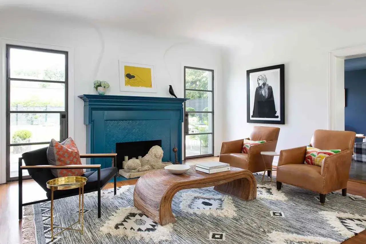 Un salon moderne avec une cheminée d'un **couleur bleu foncé** saisissante, deux fauteuils en cuir marron, un tapis graphique et une table basse en rotin courbée.