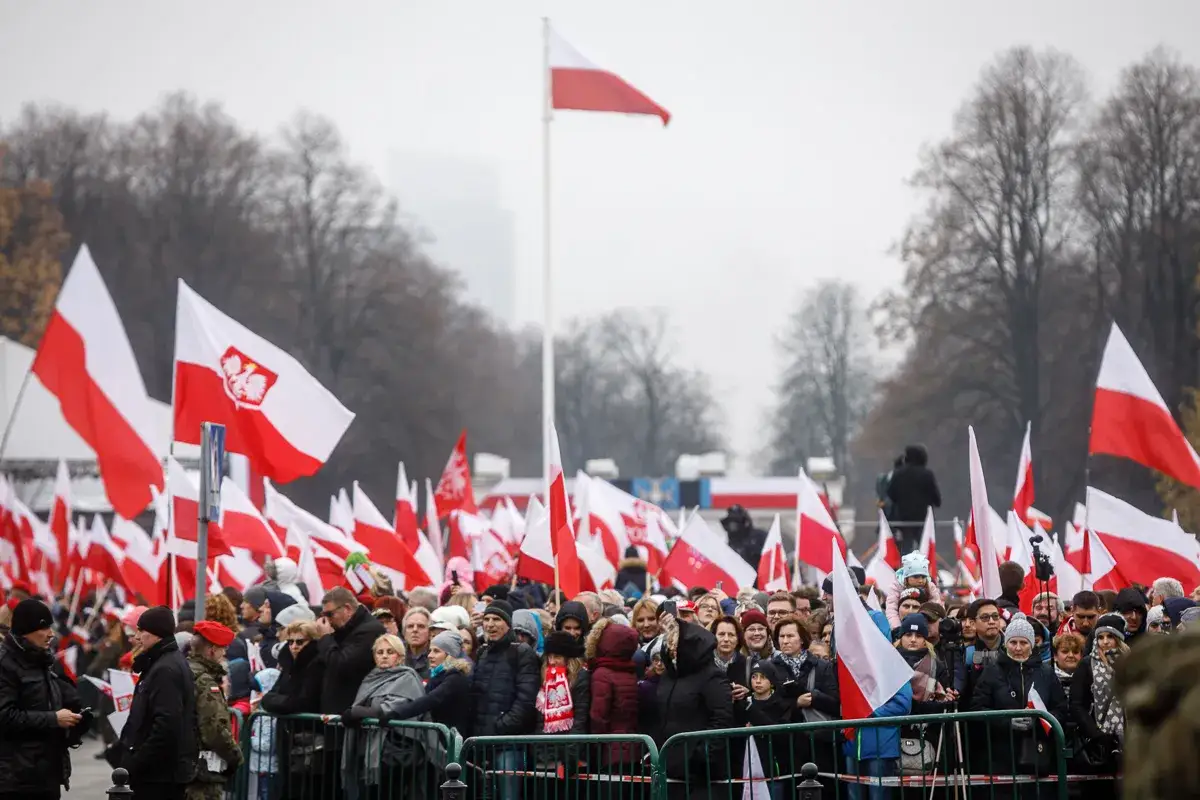 Tłum ludzi świętuje dzień niepodległości, machając polskimi flagami. Biało-czerwone barwy dominują na tle drzew i pochmurnego nieba.