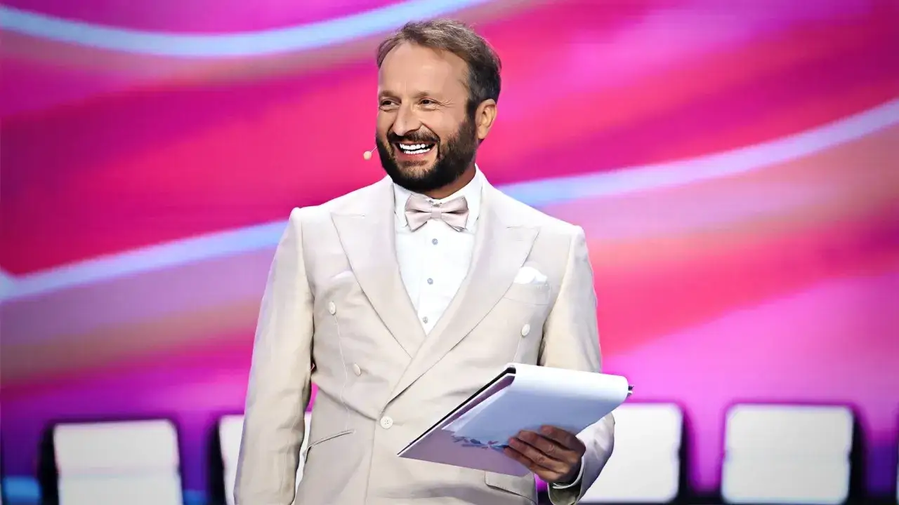Uśmiechnięty komik w beżowym garniturze trzyma dokumenty. Dowiedz się, dlaczego znany komik przestał pić alkohol i jak to wpłynęło na jego żarty.