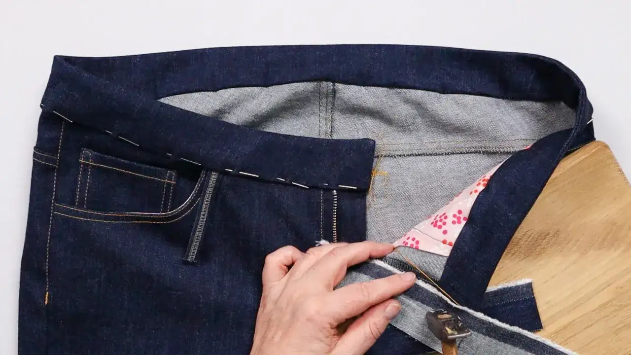 A hand uses a hammer to press down on a denim waistband, showcasing the waistband method for sewing jeans.