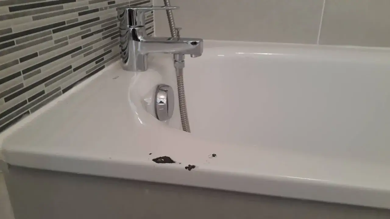Bain en &eacute;mail blanc avec une rayure sombre et des &eacute;clats sur le bord. Robinet chrom&eacute; et carrelage mural &agrave; rayures grises en arri&egrave;re-plan.