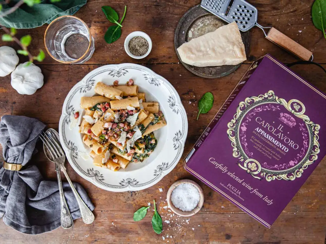 En tallrik rigatoni med pancetta och spenat, redo f&ouml;r ett gott pancetta recept. Bredvid st&aring;r en flaska vin och ingredienser.