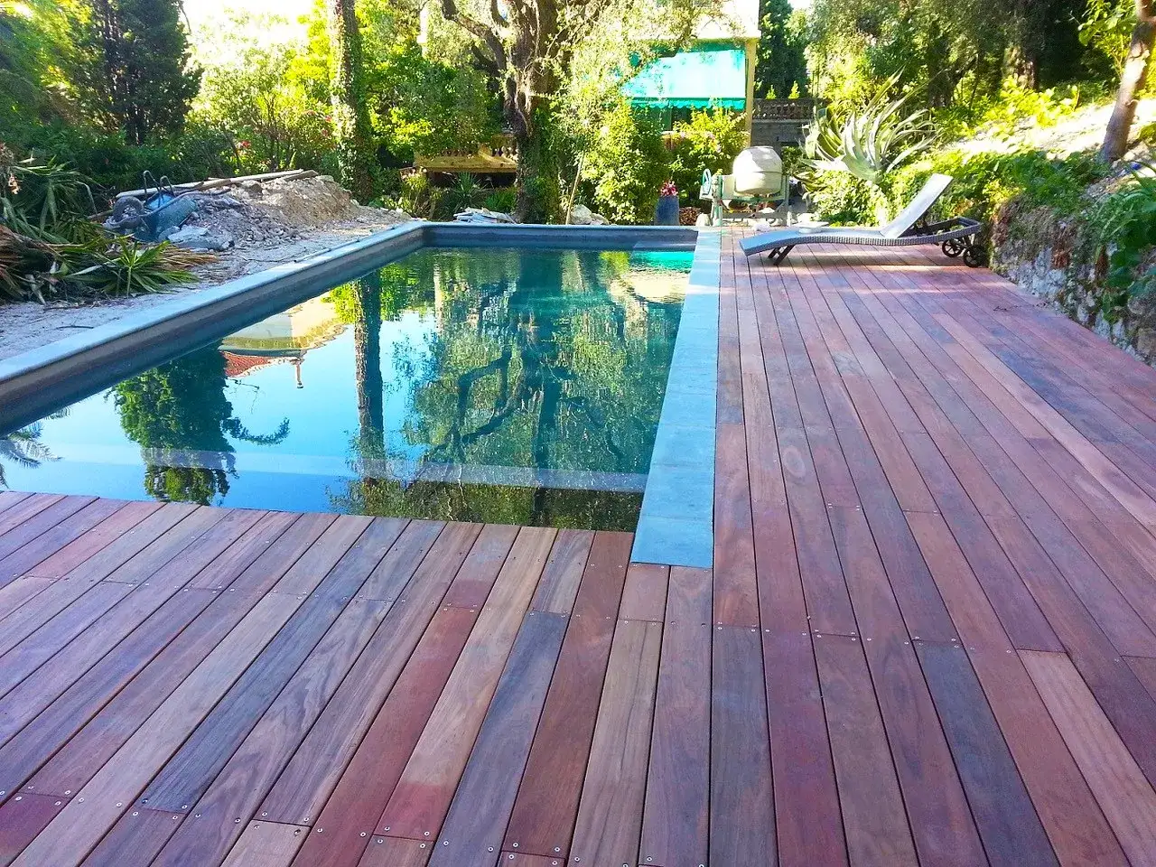 Terrasse en padouk vieillissant, refl&eacute;tant le ciel et les arbres dans la piscine. Un transat attend au soleil.