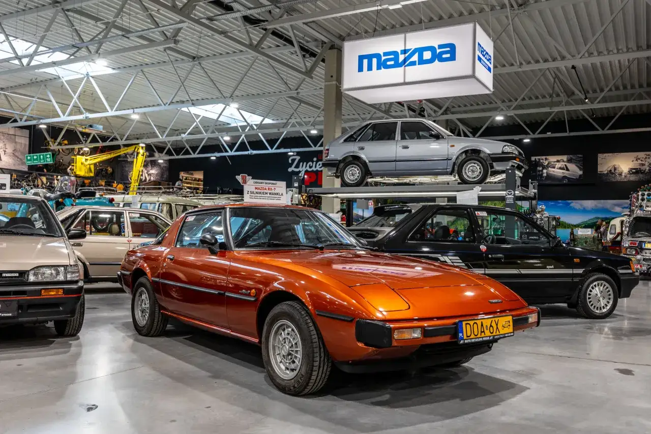 Pomarańczowa Mazda RX-7 błyszczy w muzeum samochodów, obok czarnego modelu i innych klasyków.