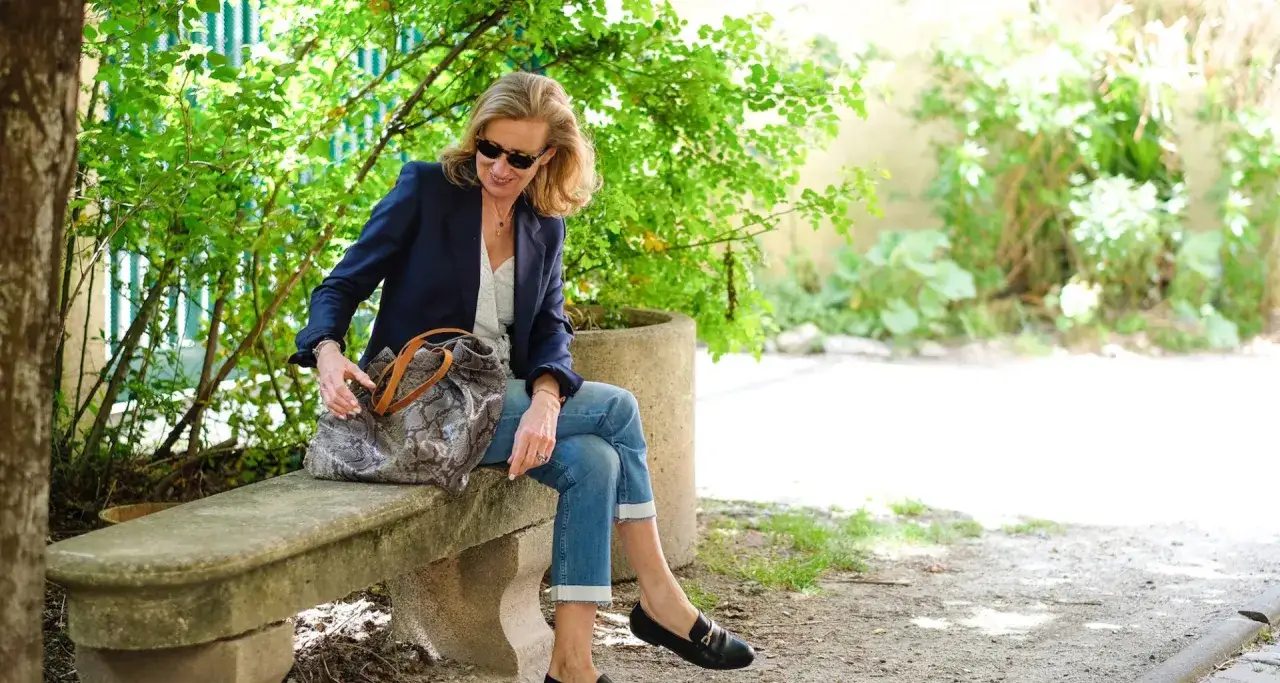 Femme assise sur un banc, portant un blazer bleu marine, un jean retroussé et un mocassin noir. Elle tient un sac à main imprimé serpent.