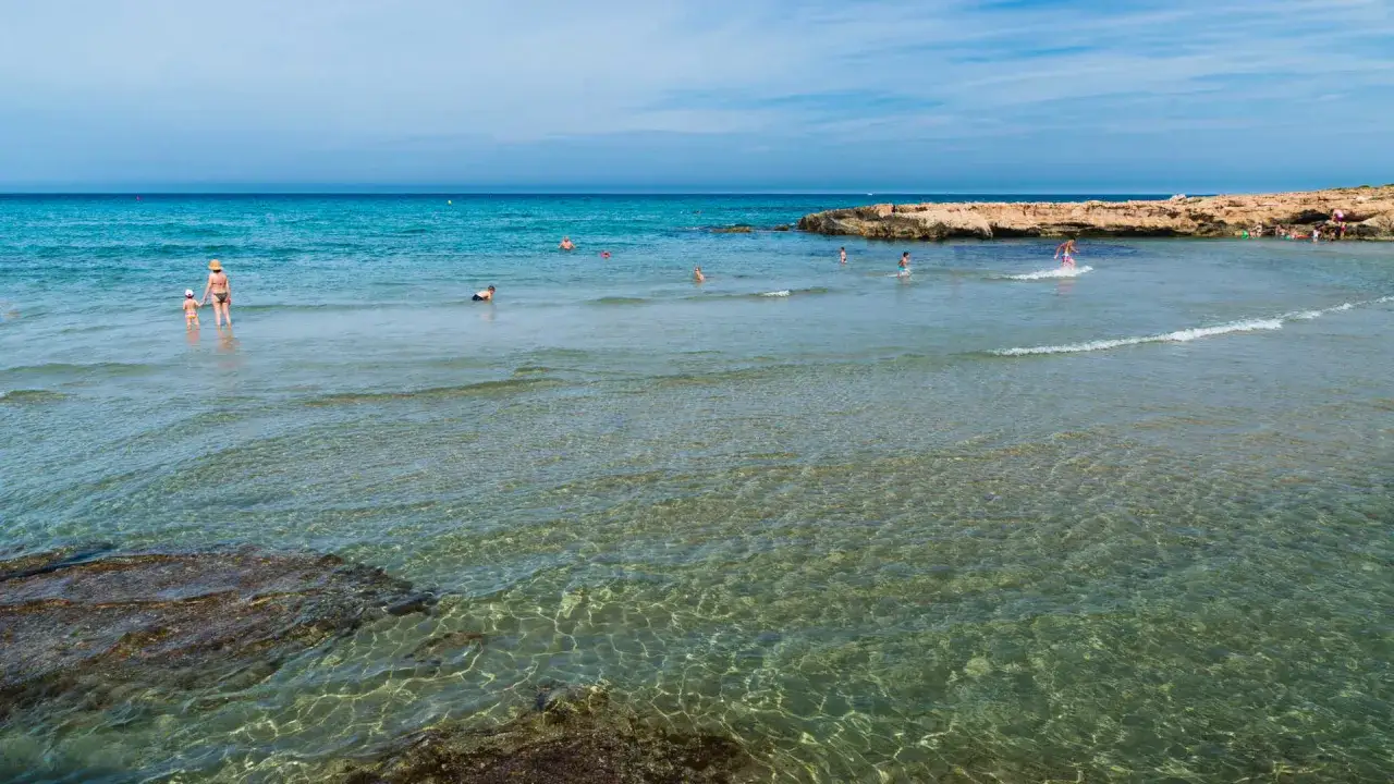 Gente disfrutando del agua cristalina en cala Capitán, con rocas y el mar azul de fondo.