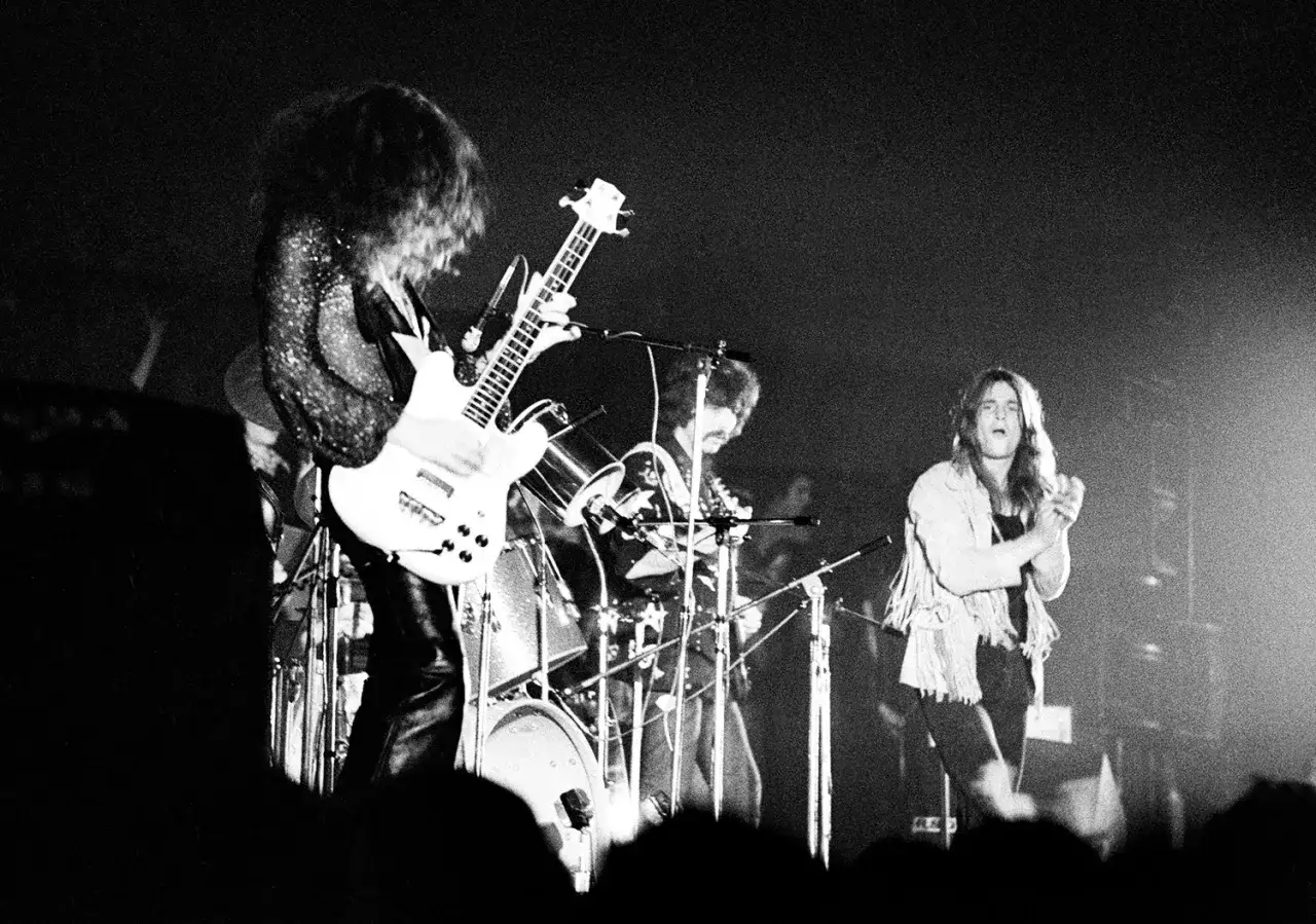 Koncert Black Sabbath, zespół gra swoje największe przeboje. Wokalista Ozzy Osbourne w centrum, gitarzysta i perkusista w tle.
