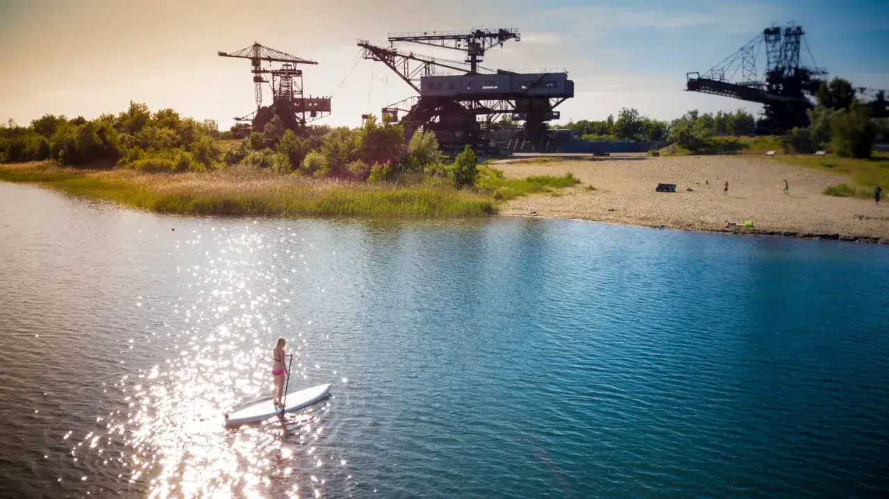 Eine Frau auf einem Stand-Up-Paddleboard genie&szlig;t die Sonne auf einem der sch&ouml;nen Badeseen Sachsen-Anhalts, w&auml;hrend im Hintergrund alte Industriekr&auml;ne stehen.