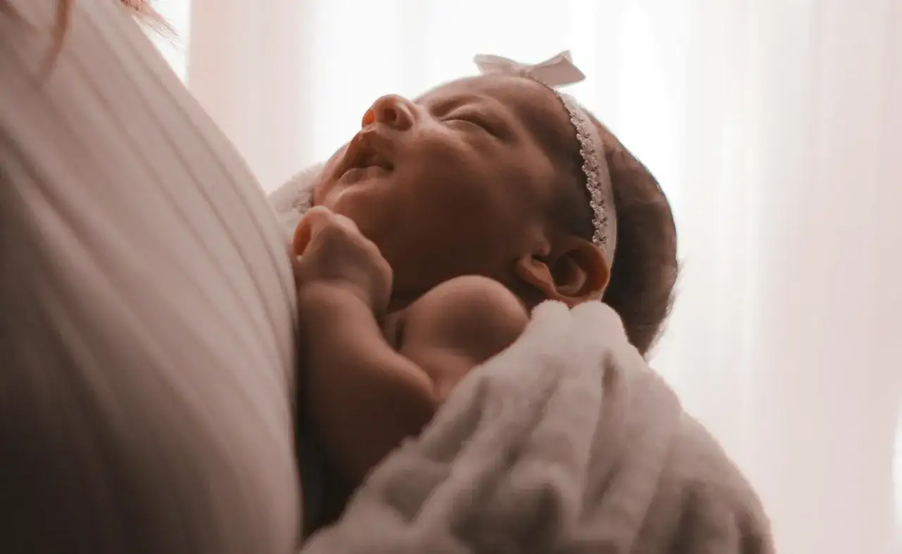 Un neonato avvolto in una coperta bianca, con un fiocco rosa in testa. I suoi piccoli tremori sono quasi impercettibili mentre dorme serenamente.