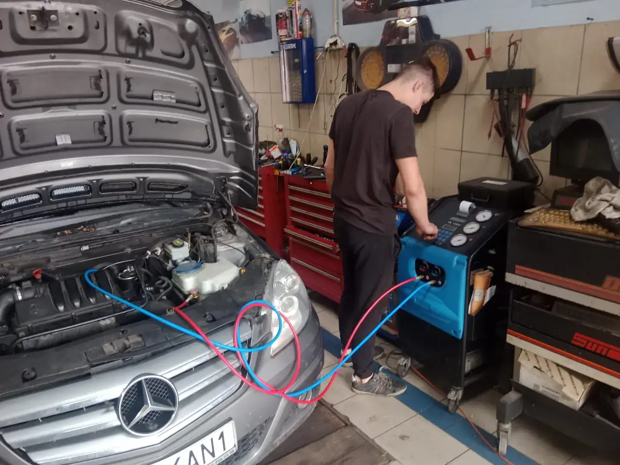 Mechanik przeprowadza nabijanie klimatyzacji w samochodzie Mercedes. Widać przewody podłączone do silnika i maszyny.