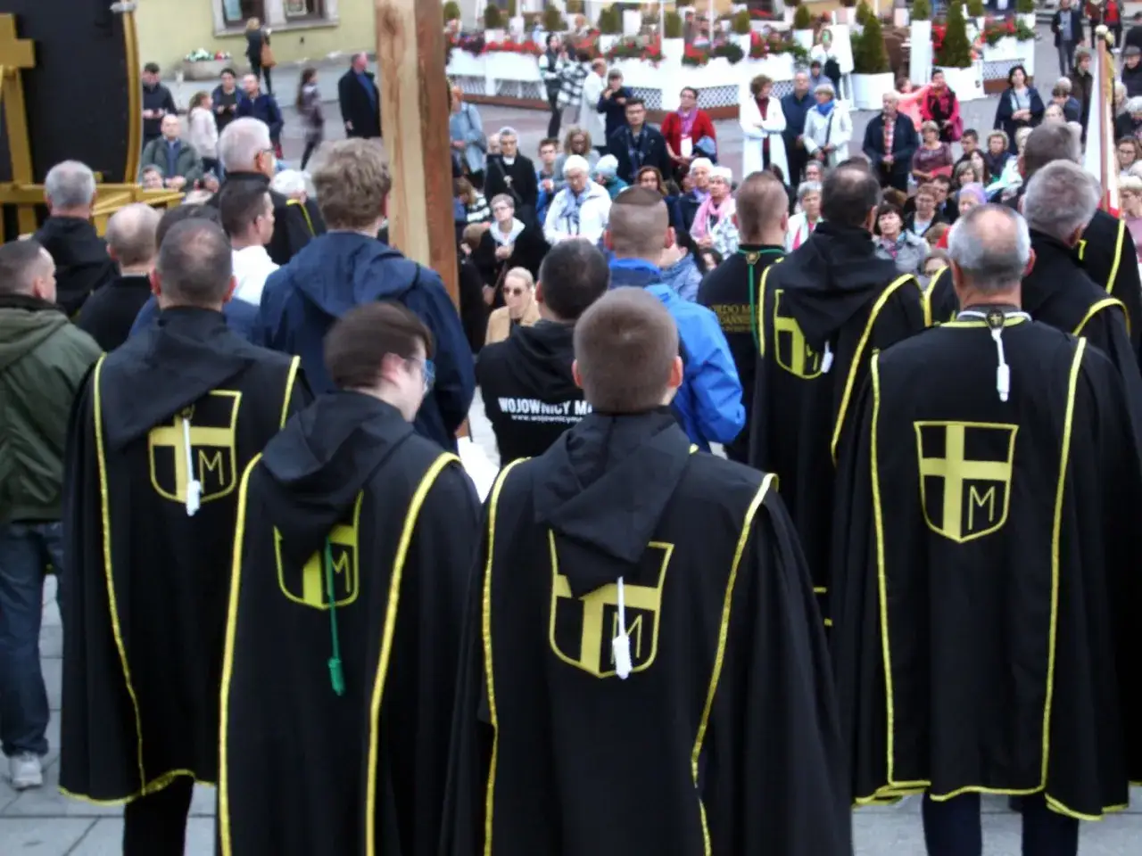Grupa mężczyzn w czarnych płaszczach z żółtymi symbolami krzyża i litery M, identyfikująca się jako rycerze Maryi, uczestniczy w uroczystości.