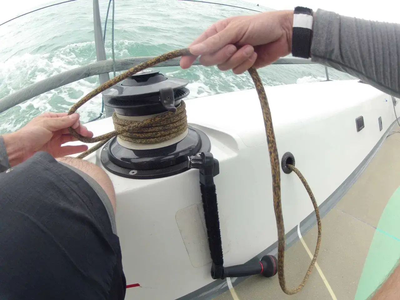 Mains de marin manœuvrant une corde sur un winch, prêt à embraquer voile. L'eau clapote, le vent souffle.