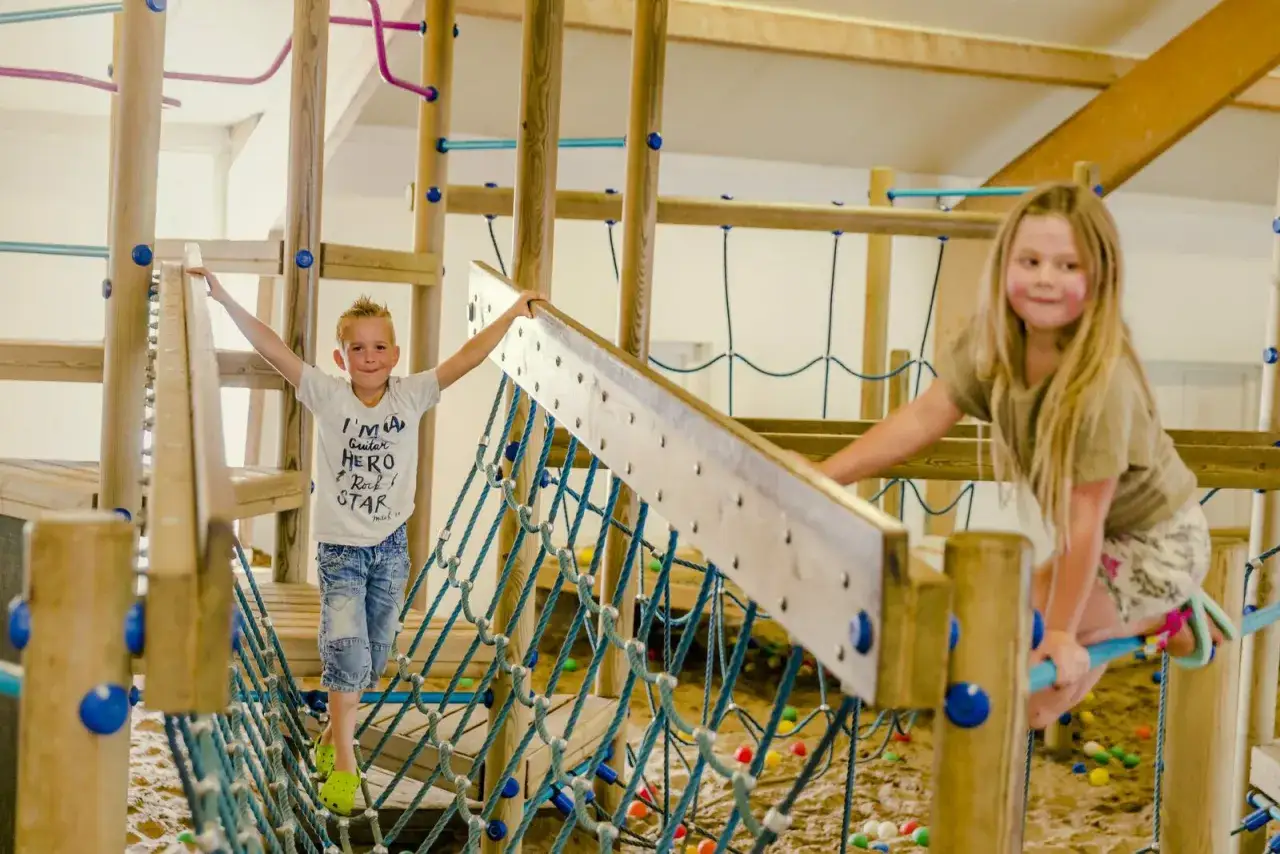Zwei Kinder balancieren auf einer H&auml;ngebr&uuml;cke in einem Indoor-Spielplatz. Ein tolles Abenteuer, fast wie beim Zelten mit Kindern!