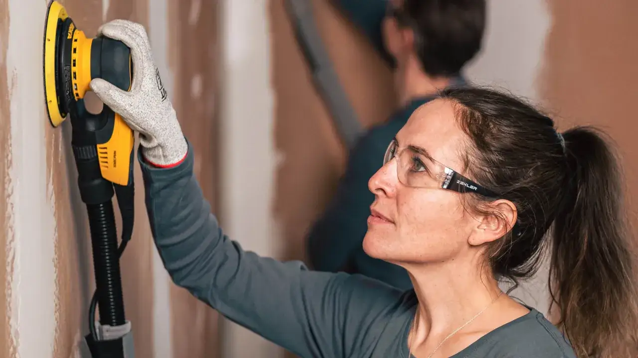 Donna con occhiali protettivi usa una levigatrice per imparare come levigare un muro.