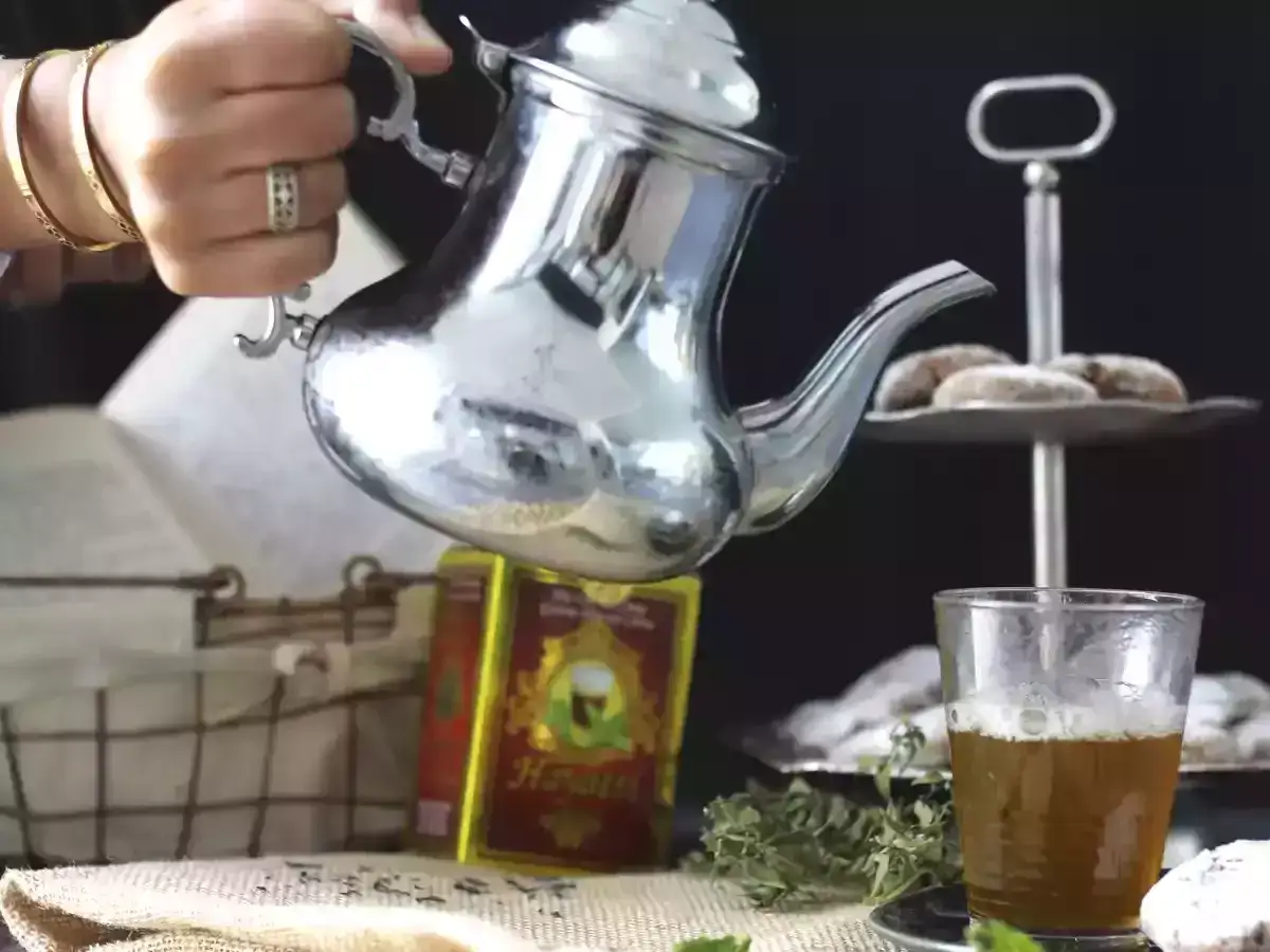 Ręka nalewa aromatyczną herbatę marokańską z ozdobnego czajniczka do szklanki. Obok ciasteczka i świeże zioła.