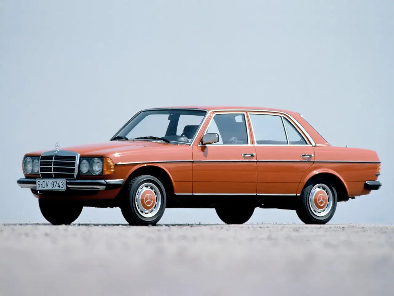 En orange Mercedes 60-tal, en klassisk sedan med kromdetaljer, står parkerad på en grusväg mot en ljusblå himmel.