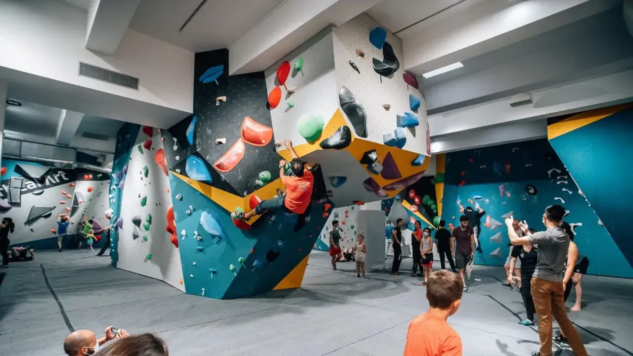 Un homme en t-shirt orange pratique l'escalade sur un mur coloré, entouré d'autres grimpeurs et spectateurs dans une salle d'escalade. C'est un vrai centre d'escalade nation !