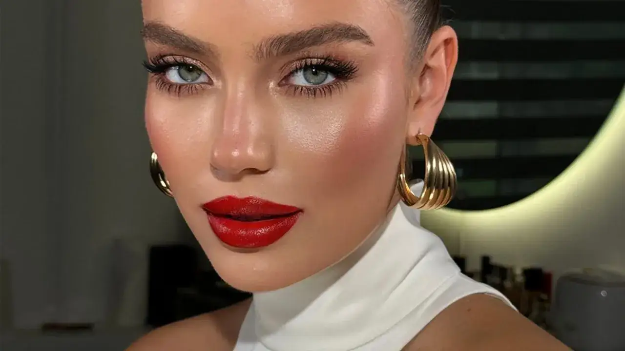 Primo piano di una donna con un trucco impeccabile, labbra definite da un audace rossetto rosso e grandi orecchini dorati.