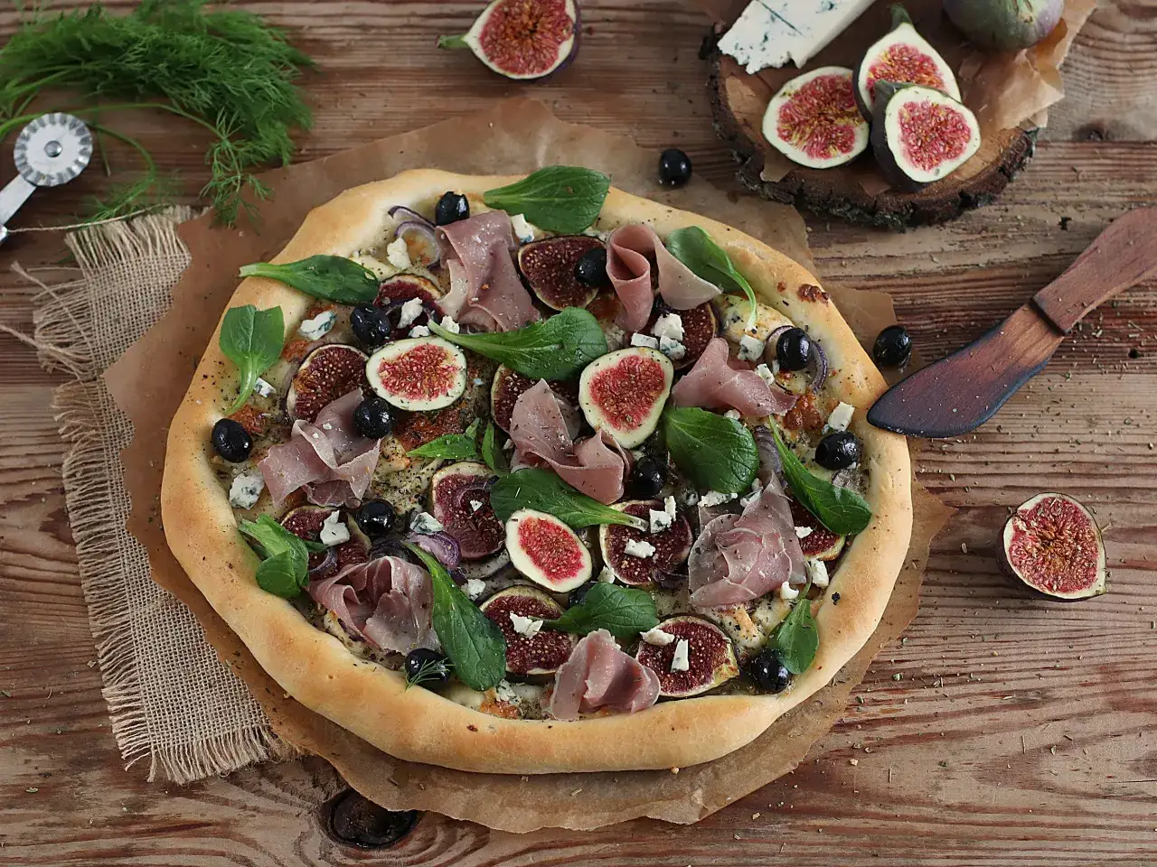 Pizza z figami, szynką parmeńską, oliwkami i serem pleśniowym. Widać też świeże liście szpinaku i cebulę. To idealne pizza składniki na wierzch.