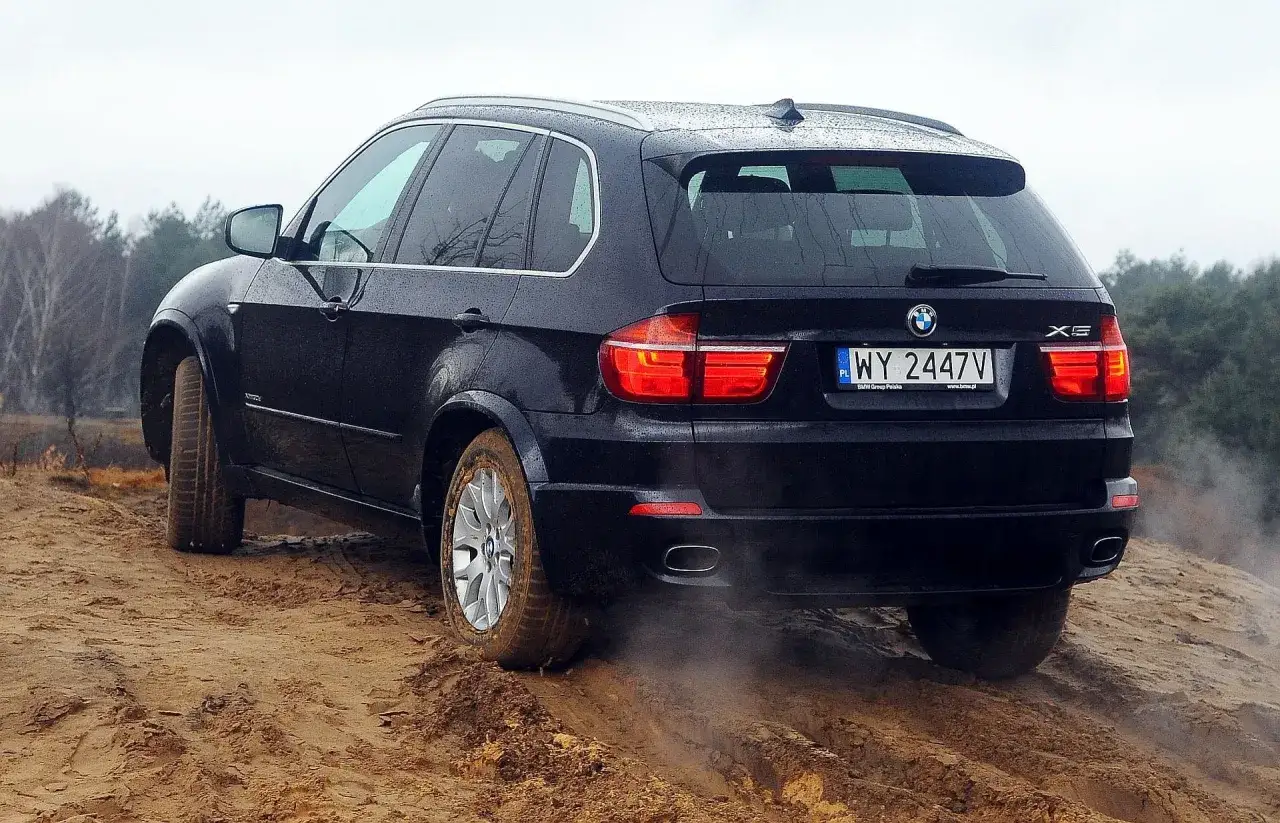 Czarne BMW X5 E70 na piaszczystym podjeździe, koła w błocie, kurz wzbija się w powietrze.