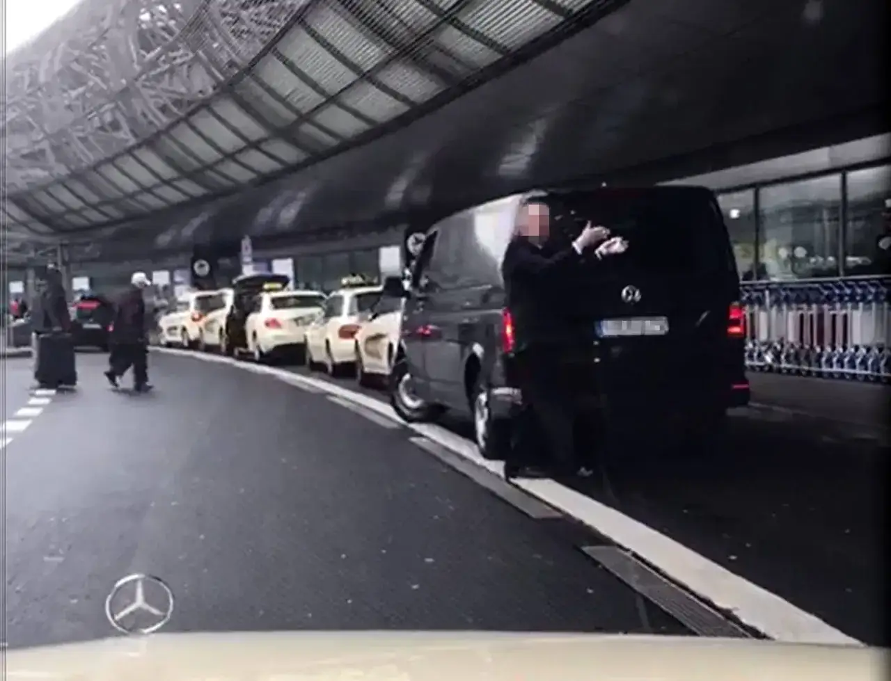 Ein Mann in Schwarz steht neben einem schwarzen Van, während Taxis am Flughafen warten. Er scheint über seine Arbeit nachzudenken.