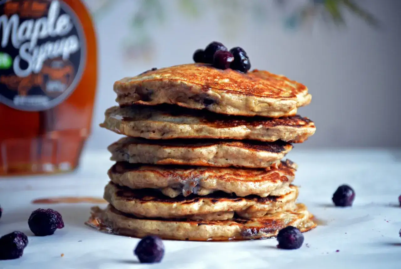 Ein Stapel zuckerfreie Pancakes mit Blaubeeren, daneben eine Flasche Ahornsirup. Ein gesunder Genuss!