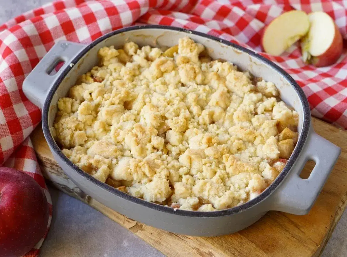 Ein goldbrauner Apfel Crumble in einer Auflaufform, bereit zum Genießen. Daneben liegen frische Äpfel auf kariertem Tuch.