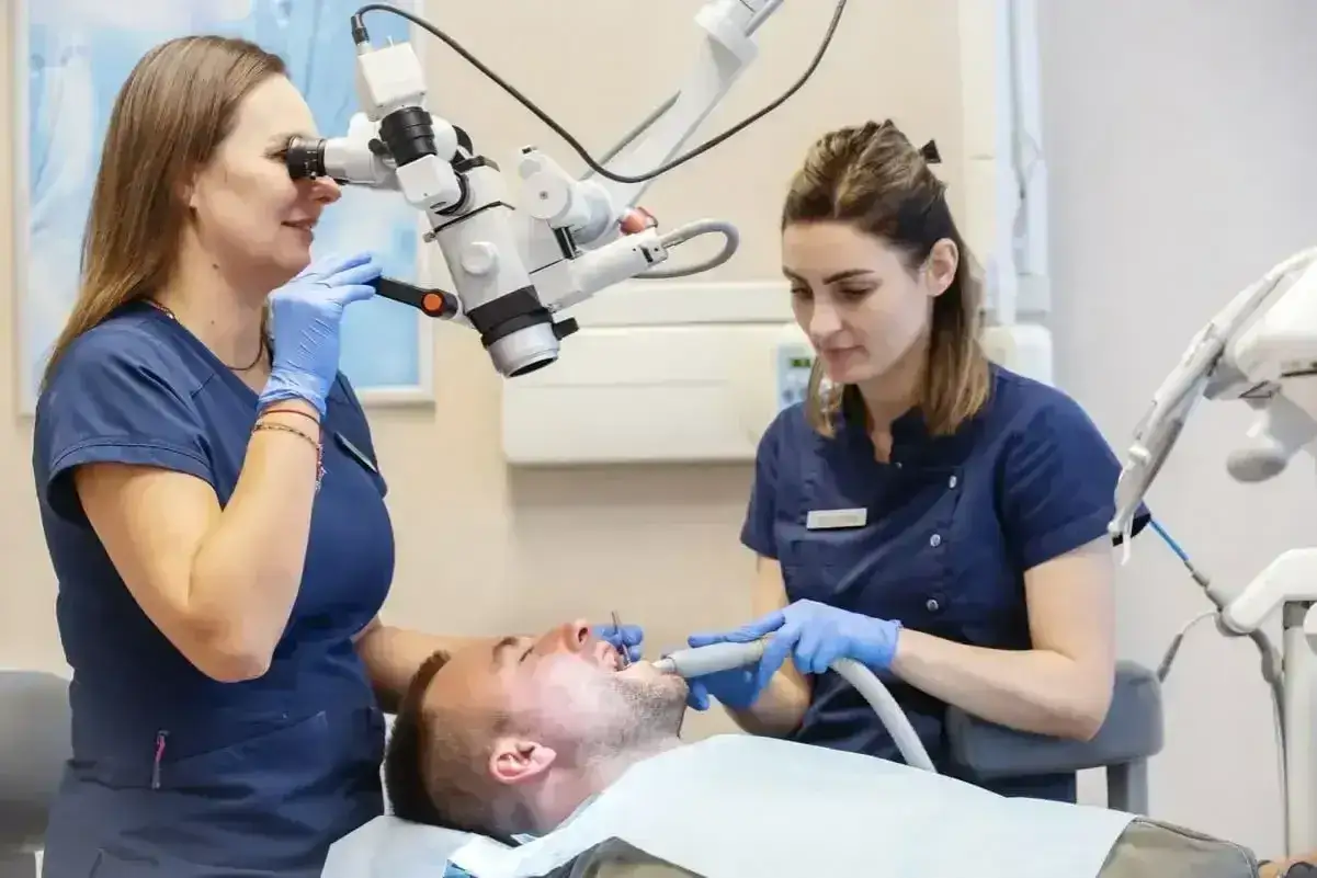 Dwie dentystki pracują przy pacjencie. Jedna używa mikroskopu, druga narzędzia. Czy znieczulenie u dentysty boli? Tu tego nie widać.