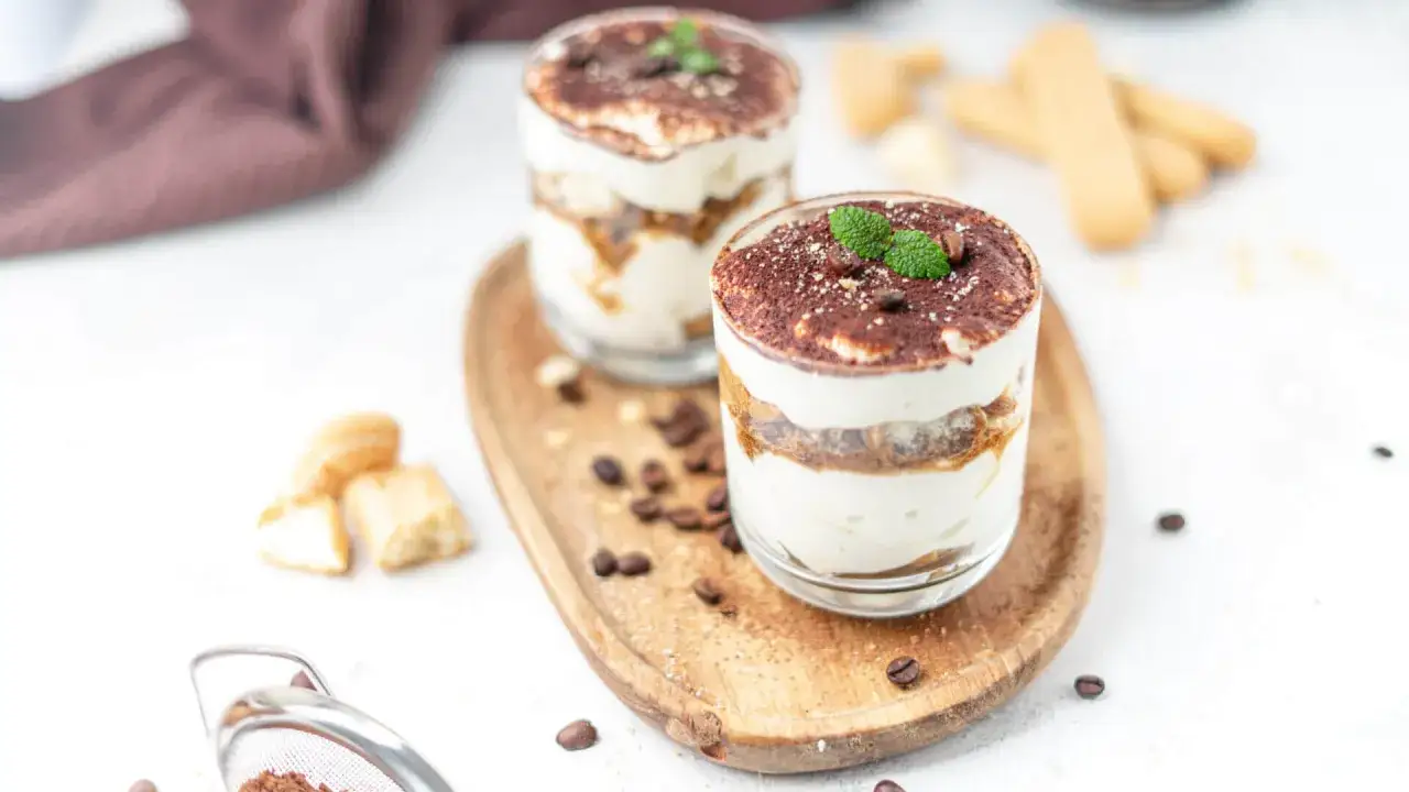 Zwei Gl&auml;ser Tiramisu ohne Zucker, garniert mit Minze und Kakaopulver, auf einem Holztablett mit Kaffeebohnen.