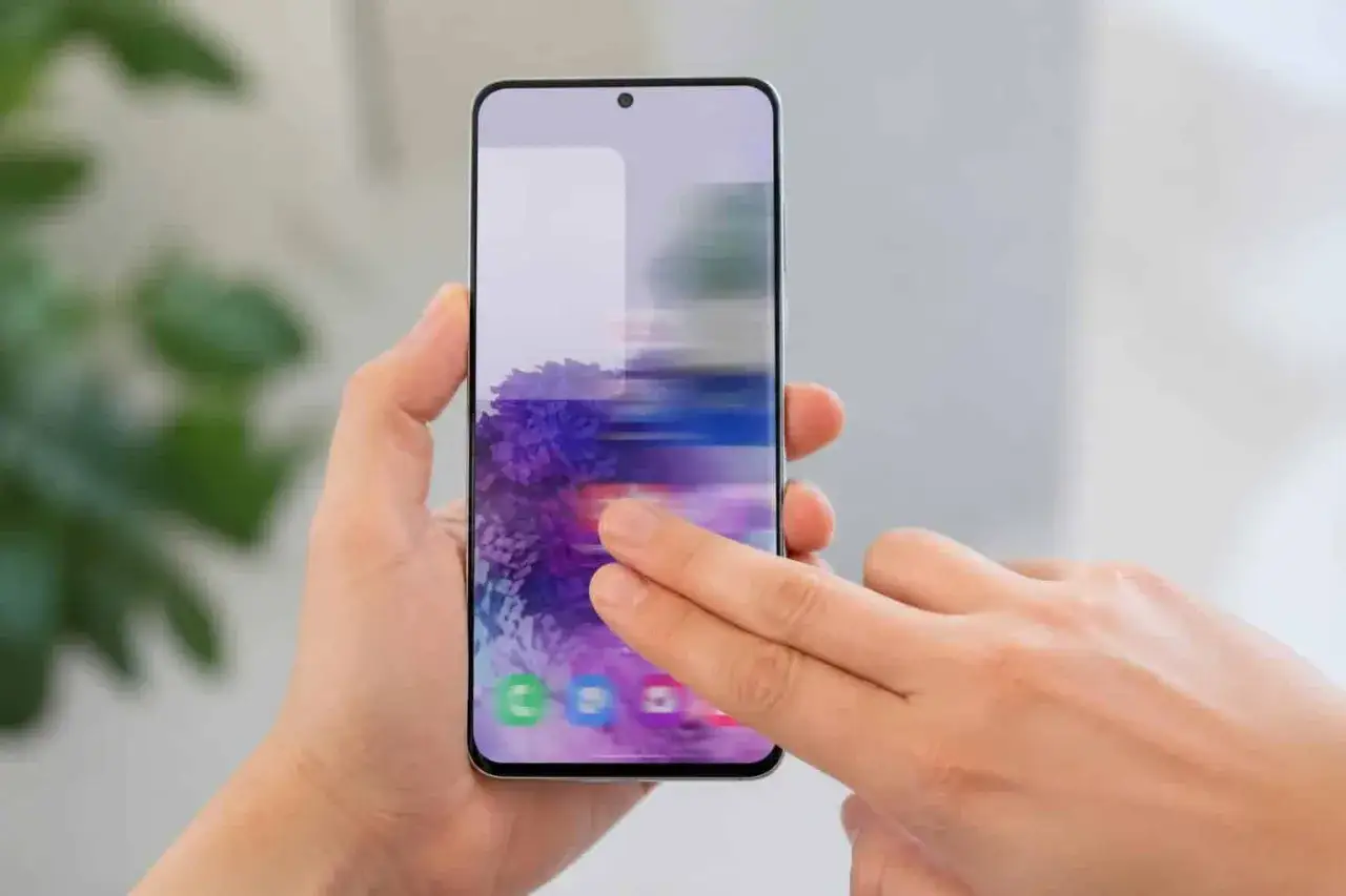 Mains tenant un smartphone, l'une d'elles fait défiler une image floue de fleurs violettes.