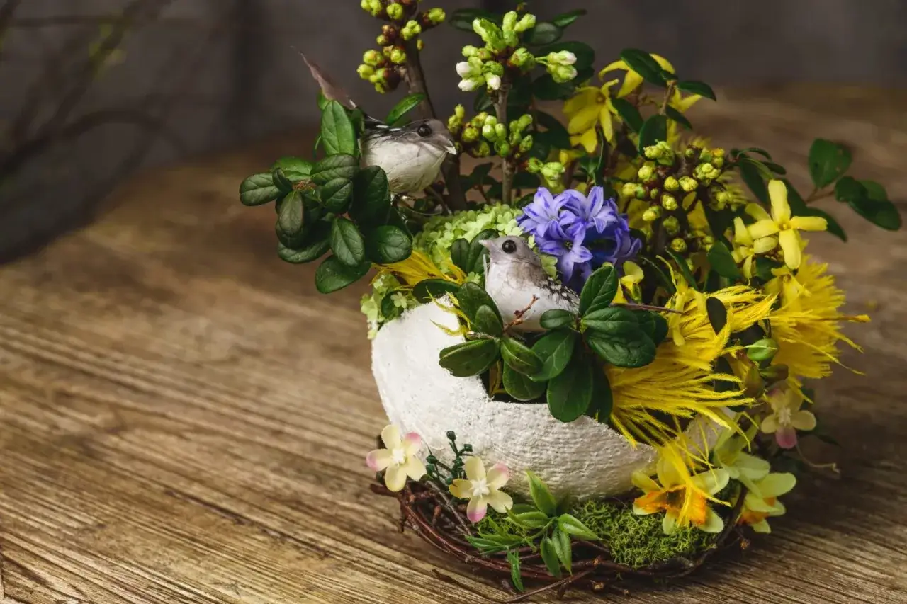 Un nid de paques composition florale de paques avec deux oiseaux, des fleurs jaunes et bleues, et des branches vertes sur un fond en bois.
