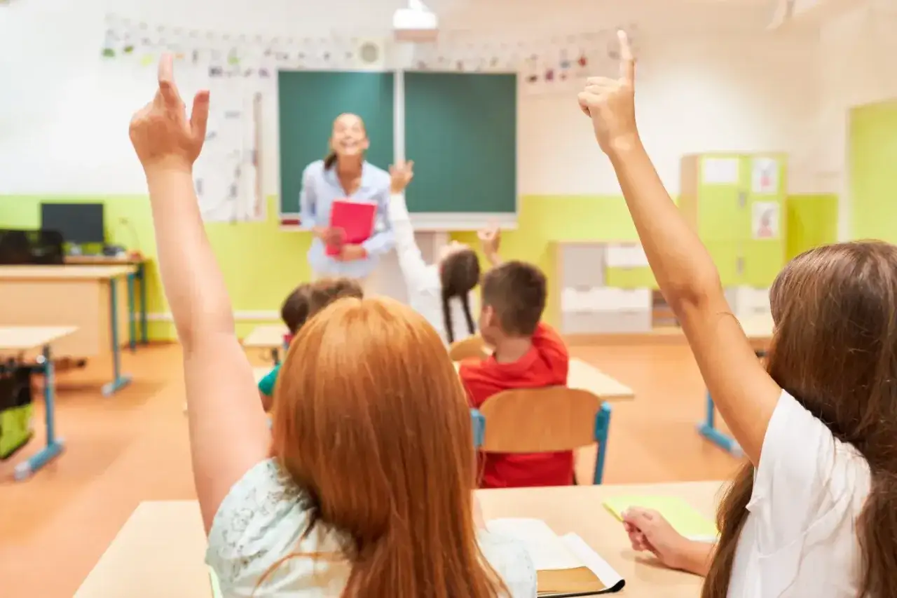Uczniowie z podniesionymi rękami, gotowi do odpowiedzi, celebrują Dzień Edukacji Narodowej. Nauczycielka z uśmiechem czeka na zgłoszenia.