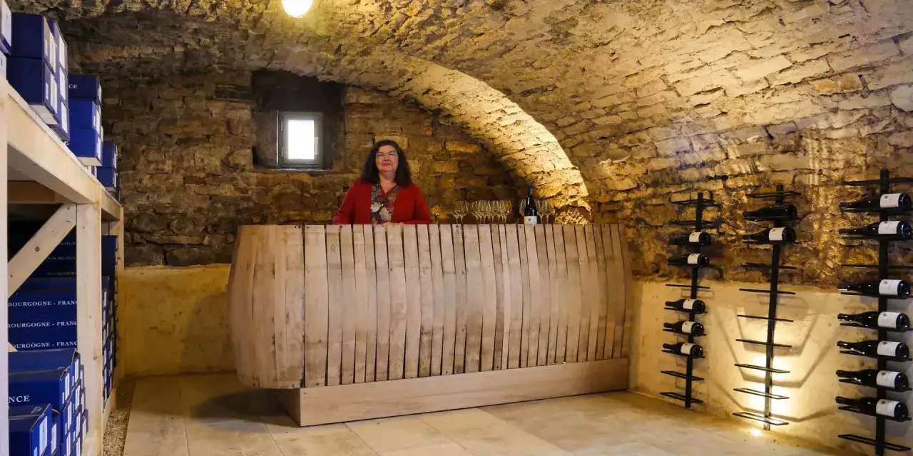Une femme accueille les visiteurs pour une visite cave Beaune. Des bouteilles de vin sont rangées sur des étagères murales.