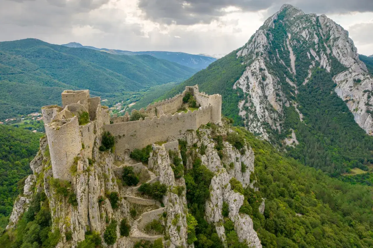 Vestiges d'un **ch&acirc;teau abandonn&eacute; en Occitanie**, perch&eacute; sur une falaise rocheuse, dominant des vall&eacute;es verdoyantes sous un ciel orageux.