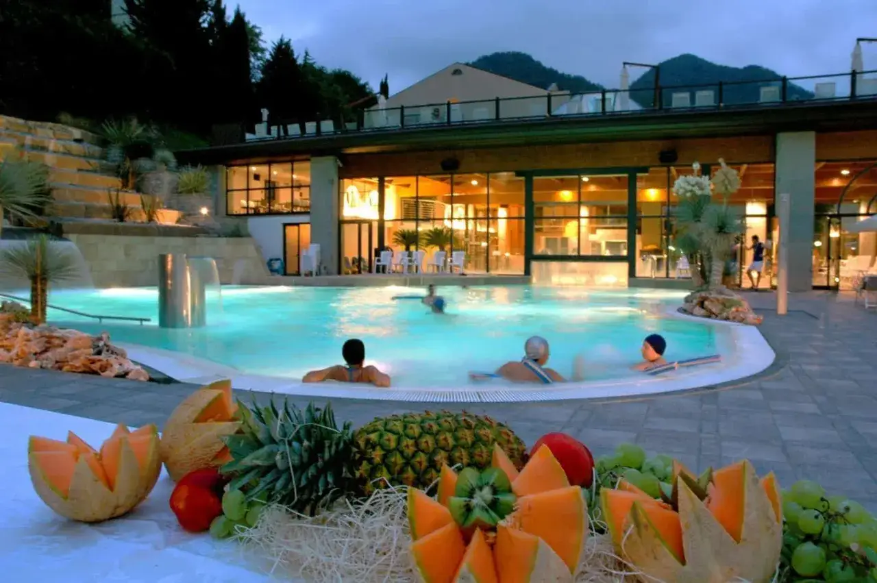 Piscina termale illuminata di notte, con persone che nuotano e un tavolo di frutta fresca in primo piano. Un'oasi di relax alle terme di Reggio Emilia.