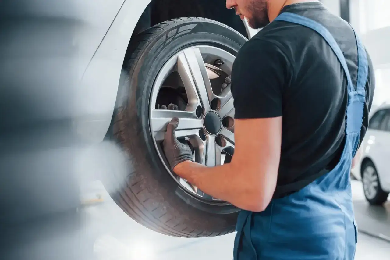 Mechanik w niebieskim kombinezonie sprawdza budowę koła samochodowego. W tle biały samochód i unosząca się mgiełka.